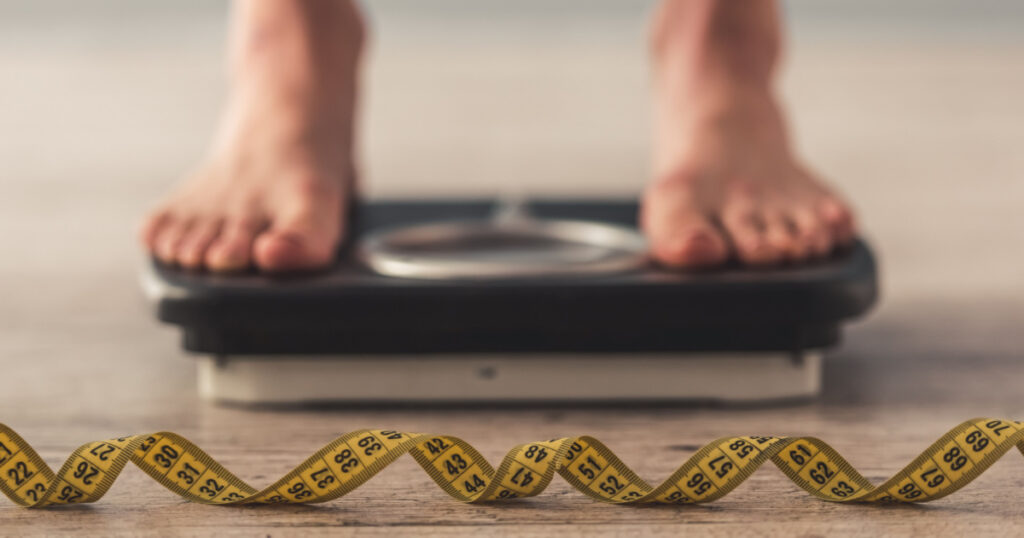 Cropped image of woman feet standing on weigh scales, on gray background. A tape measure in the foreground